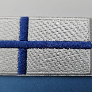 Op de afbeelding: Geborduurde patch met de Finse vlag. De rechthoekige patch heeft een witte achtergrond met een blauw Scandinavisch kruis. Het kruis loopt door tot aan de randen van de patch. De patch is waarschijnlijk voor kleding of accessoires.