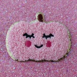 Op de afbeelding: Een roze, pluche pompoenpatch met een lachend gezicht, zwarte wimpers en roze wangen. De pompoen heeft een gouden rand en een bruine steel. De achtergrond is een sprankelend roze.