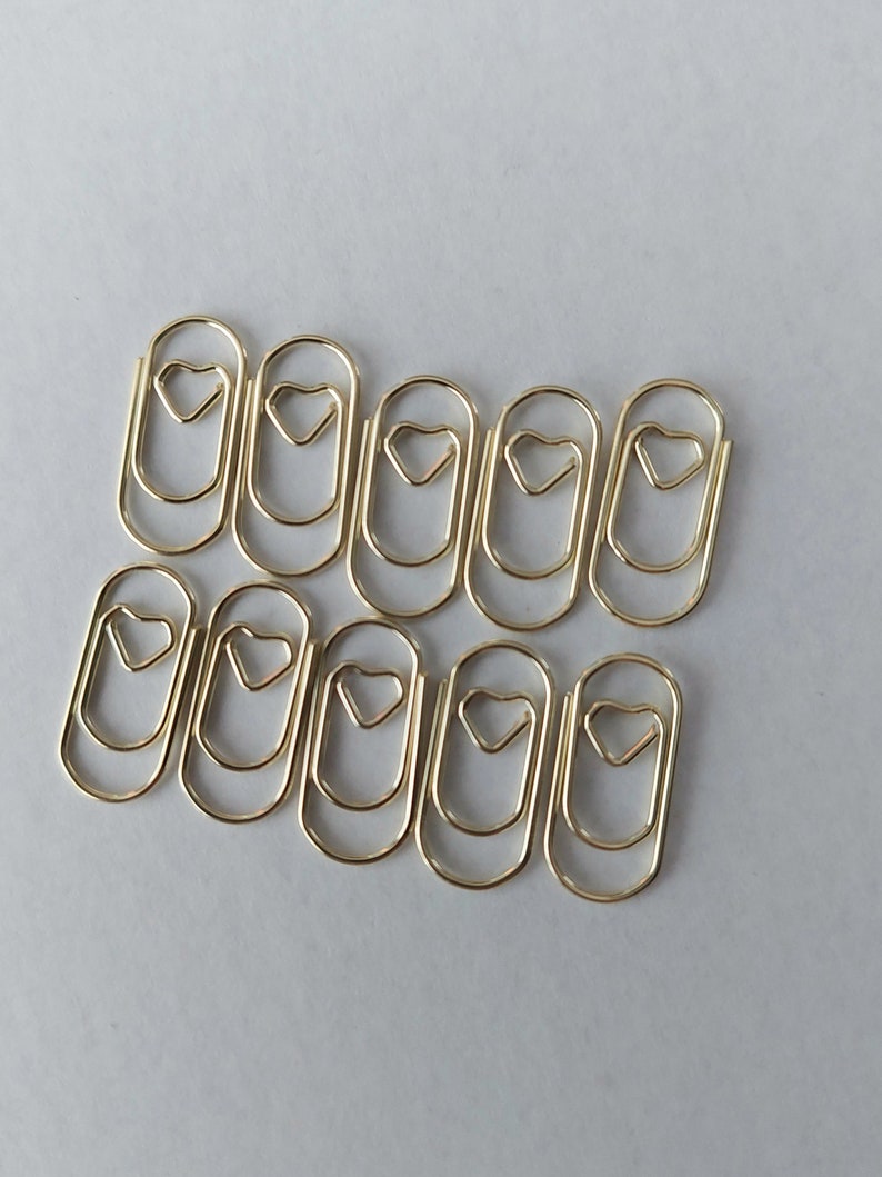 K&ouml;nnte beinhalten: Zehn goldfarbene B&uuml;roklammern mit herzf&ouml;rmigem Design. Die Clips liegen auf einer wei&szlig;en Oberfl&auml;che und eignen sich als dekoratives und praktisches B&uuml;roaccessoire.