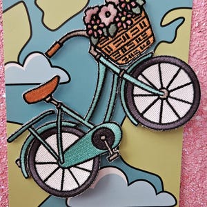 May include: Embroidered patch depicting a teal bicycle carrying a basket of pink and purple flowers. The bicycle has white wheels and a brown seat. The patch is on a blue and green background with the text "DIAMONDHEARTNL".