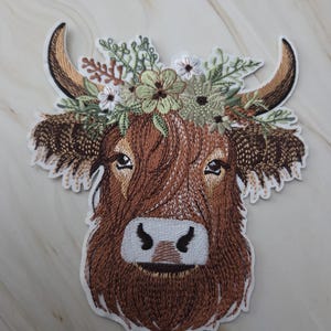 Op de afbeelding: Geborduurde patch met een Schotse Hooglander met bruine vacht, hoorns en een bloemenkrans. De patch heeft een witte rand en gedetailleerde stiksels, die het gezicht van de koe en het bloemendesign laten zien.