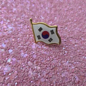 Puede incluir: Un pin con borde dorado que representa la bandera de Corea del Sur, con un fondo blanco, un símbolo Taegeuk rojo y azul y cuatro trigramas negros. El pin está sobre un fondo rosa brillante.