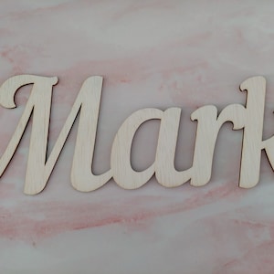 Op de afbeelding: Houten uitsparing van de naam "Mark" in een cursief lettertype. De letters zijn licht van kleur en lijken van onbewerkt hout te zijn. De achtergrond is een roze en wit marmerpatroon.