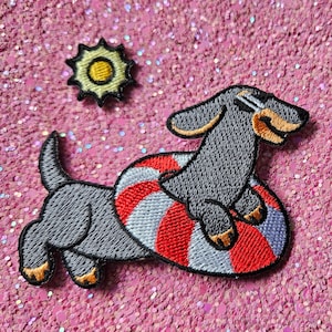 Op de afbeelding: Geborduurde patch met een grijze teckel met een zonnebril en een rood, wit en blauw gestreepte zwemband. Er is ook een kleine zon-patch inbegrepen. De patches staan op een roze glitterachtergrond.