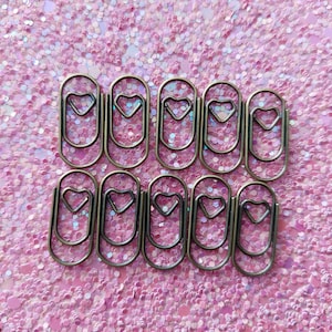 K&ouml;nnte beinhalten: Zehn metallene B&uuml;roklammern, jede mit einem herzf&ouml;rmigen Design, liegen auf einem rosa Glitzerhintergrund. Diese dekorativen Klammern verleihen Schreibwaren eine charmante Note und sind ideal zum Ordnen von Papieren.
