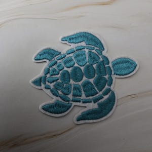 Op de afbeelding: Geborduurde turquoise zeeschildpad patch met een witte rand. De schildpadschild heeft een gedetailleerd patroon en de patch is ontworpen voor toepassing op kleding of accessoires. Een decoratief element voor knutselprojecten.