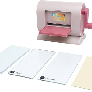 Puede incluir: Máquina de troquelado y relieve rosa y blanca con manivela. Incluye placas acrílicas transparentes etiquetadas como "Base", "Cutting" y "Embossing", además de una placa amarilla. La máquina tiene una bandeja y un asa rosas. La palabra "Mini" es visible.