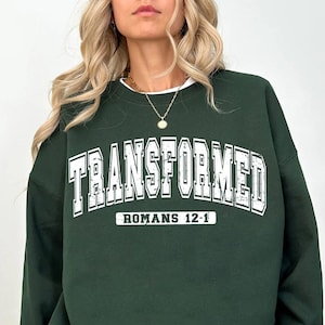 May include: A dark green sweatshirt with the text "TRANSFORMED" in white with a white outline. Below the text is "ROMANS 12-1" in white.