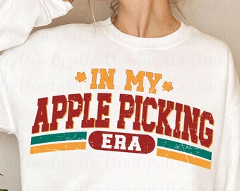 May include: White sweatshirt with a red, green, and yellow graphic that says "In My Apple Picking Era".