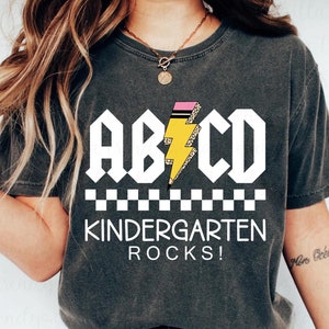 May include: Dark gray t-shirt with the text "ABCD Kindergarten Rocks!" in white block letters. A yellow lightning bolt with a pencil design is in the center. The shirt has a checkerboard pattern at the bottom.