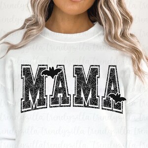 May include: A white sweatshirt with the word "MAMA" printed in black with a spiderweb pattern and bats on either side.