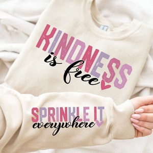 以下が含まれることがあります： ピンク、パープル、ホワイトの「Kindness is free」の文字と小さなピンクのハートがついたベージュのトレーナー。トレーナーの袖には、ピンク、パープル、ホワイトの「Sprinkle it everywhere」の文字と小さなピンクのハートが描かれています。