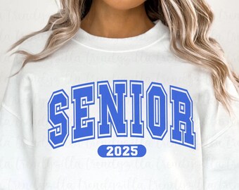 Könnte beinhalten: Weißes Sweatshirt mit dem Wort "SENIOR" in kräftigen blauen Buchstaben, weiß umrandet. Unter dem Wort "SENIOR" steht die Zahl "2025" in Blau. Das Sweatshirt hat einen Rundhalsausschnitt.