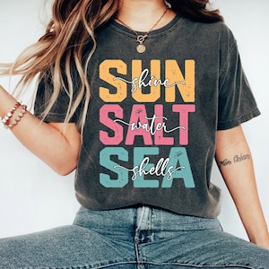 May include: A dark gray t-shirt with the words "SUN shine SALT water SEA shells" printed in colorful block letters.