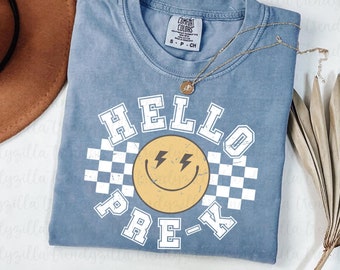 May include: A light blue t-shirt with a white graphic design that says "HELLO PRE-K" with a yellow smiley face with lightning bolts for eyes and a checkered pattern.