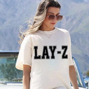May include: Cream-colored t-shirt with the text "LAY-Z" in black block letters. The shirt is worn by a person leaning against a vintage white car. The background features a mountain range and blue sky.
