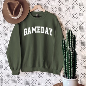 K&ouml;nnte beinhalten: Ein gr&uuml;ner Pullover mit dem Wort "GAMEDAY" in wei&szlig; auf der Vorderseite. Der Pullover h&auml;ngt an einem Kleiderb&uuml;gel vor einem wei&szlig;en Hintergrund mit einem schwarzen geometrischen Muster.