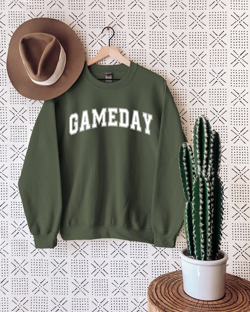 K&ouml;nnte beinhalten: Ein gr&uuml;ner Pullover mit dem Wort "GAMEDAY" in wei&szlig; auf der Vorderseite. Der Pullover h&auml;ngt an einem Kleiderb&uuml;gel vor einem wei&szlig;en Hintergrund mit einem schwarzen geometrischen Muster.