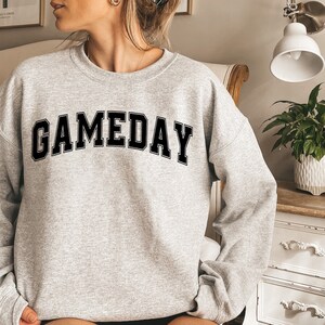 K&ouml;nnte beinhalten: Ein grauer Pullover mit dem Wort "GAMEDAY" in schwarzen Blockbuchstaben.