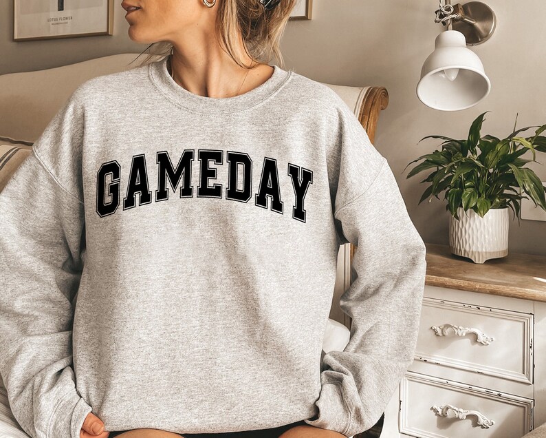 K&ouml;nnte beinhalten: Ein grauer Pullover mit dem Wort "GAMEDAY" in schwarzen Blockbuchstaben.