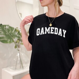 K&ouml;nnte beinhalten: Schwarzes Oversize-T-Shirt mit wei&szlig;em Text, der "GAMEDAY" in einer College-Schriftart lautet.