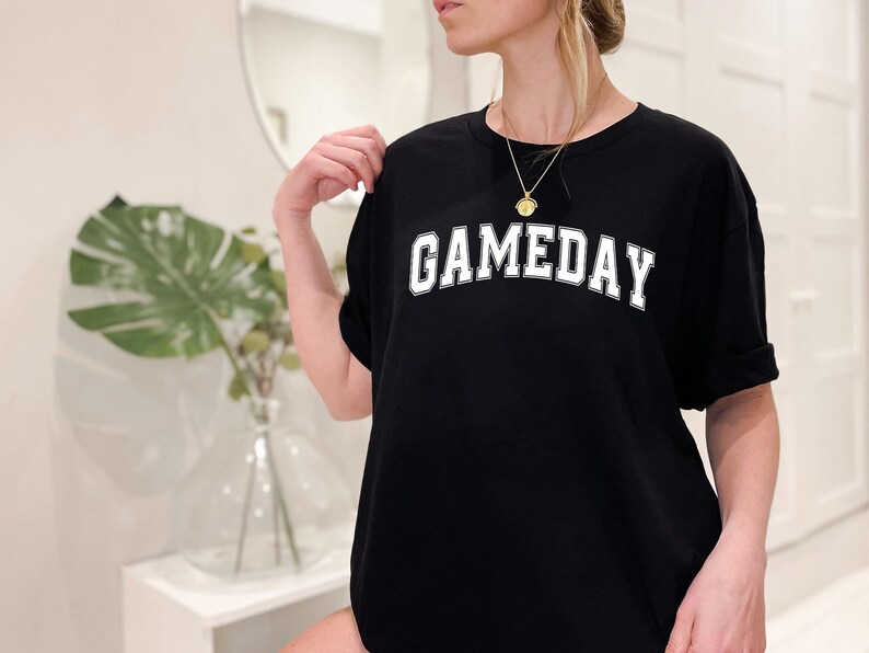 K&ouml;nnte beinhalten: Schwarzes Oversize-T-Shirt mit wei&szlig;em Text, der "GAMEDAY" in einer College-Schriftart lautet.