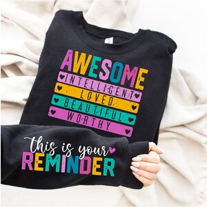 Puede incluir: Sudadera negra con el texto "AWESOME INTELLIGENT LOVED BEAUTIFUL WORTHY" en letras de colores con corazones. El texto "This is your REMINDER" está en la manga en letras de colores.