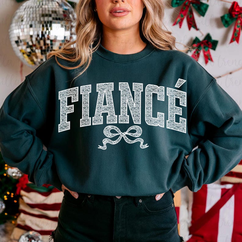 May include: A dark green sweatshirt with the word "FIANC&Eacute;" in white, outlined in a sparkly design. A white ribbon with a bow is below the word.