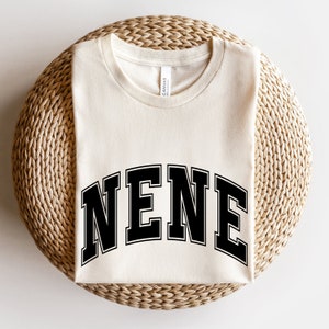 May include: Cream-colored t-shirt with the word "NENE" in bold, black, collegiate-style lettering. The shirt is folded neatly on a woven, circular surface. The shirt has a classic crew neck and short sleeves.