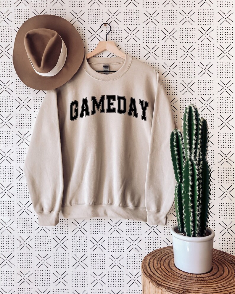 K&ouml;nnte beinhalten: Ein beiger Pullover mit dem Wort "GAMEDAY" in Schwarz auf der Vorderseite. Der Pullover h&auml;ngt an einem Kleiderb&uuml;gel vor einem wei&szlig;en Hintergrund mit einem schwarzen geometrischen Muster.