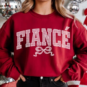 May include: Red sweatshirt with white text that says "FIANC&Eacute;" with a white bow underneath.