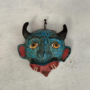 Op de afbeelding: Een blauw en turquoise masker met zwarte hoorns, gele ogen en een rode mond en wangen. Het masker hangt tegen een witte, getextureerde muur. Het masker heeft een duivels uiterlijk.