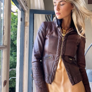 May include: A brown leather jacket with a quilted design on the shoulders and sleeves. The jacket has a black fur collar and a gold zipper. The model is wearing a gold necklace and a peach-colored dress.