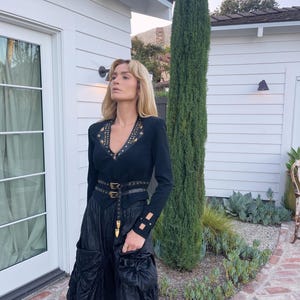 May include: A black long-sleeve top with gold-colored embellishments and a V-neck. The top is paired with a black skirt and a black belt with gold-colored buckles. The outfit is completed with a black skirt with pockets.