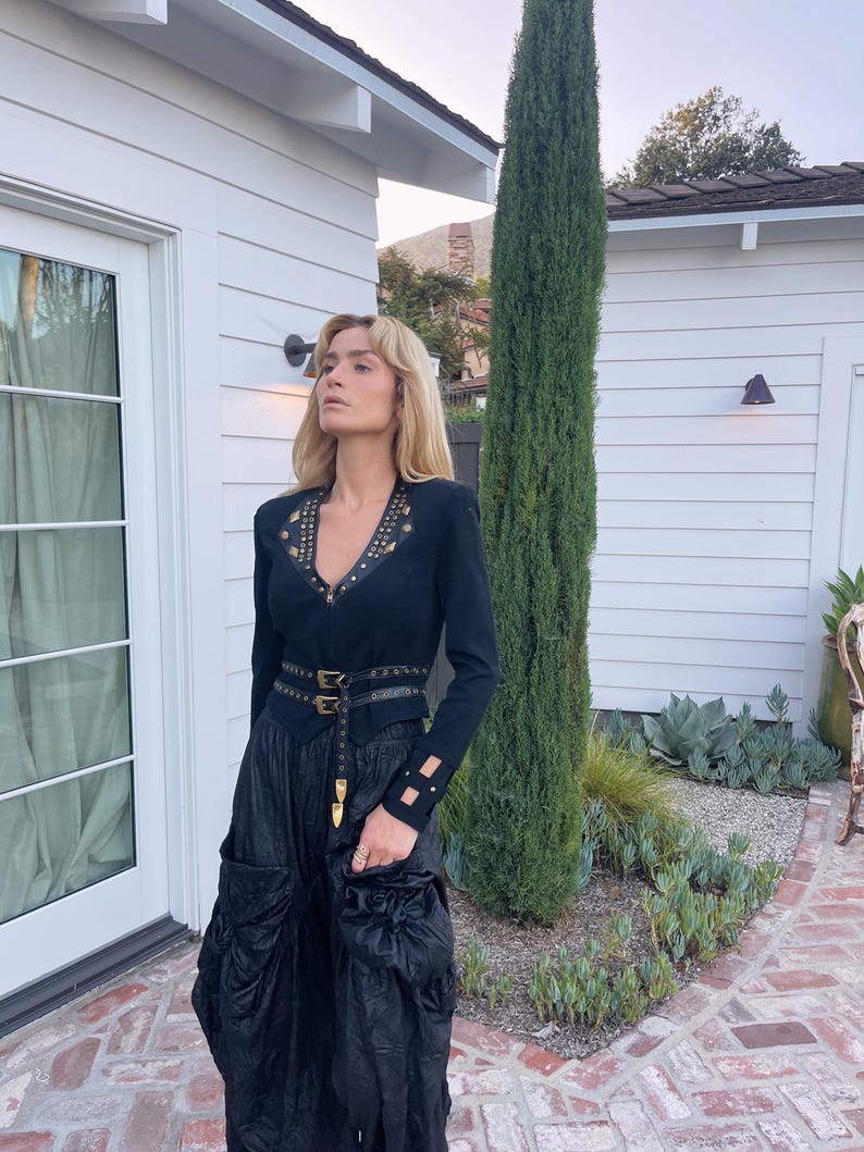 May include: A black long-sleeve top with gold-colored embellishments and a V-neck. The top is paired with a black skirt and a black belt with gold-colored buckles. The outfit is completed with a black skirt with pockets.