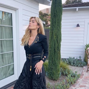 May include: A woman wearing a black outfit with a long-sleeved top and a full skirt. The top has gold-colored studs and a V-neck. A black belt with gold-colored hardware cinches the waist. The skirt has large pockets and a gathered design. The woman is standing outside.