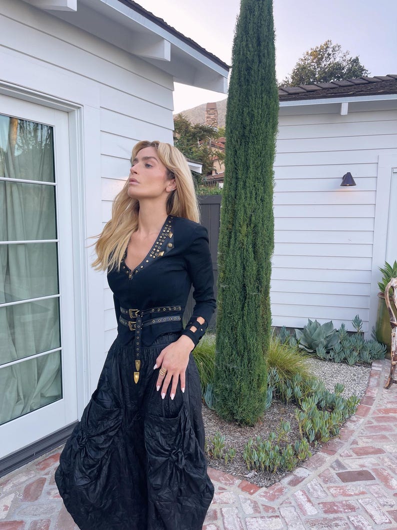 May include: A woman wearing a black outfit with a long-sleeved top and a full skirt. The top has gold-colored studs and a V-neck. A black belt with gold-colored hardware cinches the waist. The skirt has large pockets and a gathered design. The woman is standing outside.