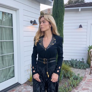 May include: Black long-sleeve top with gold-colored studs and a matching belt. The top has a zipper closure and cut-out details on the sleeves. The outfit is paired with black, wide-leg pants.