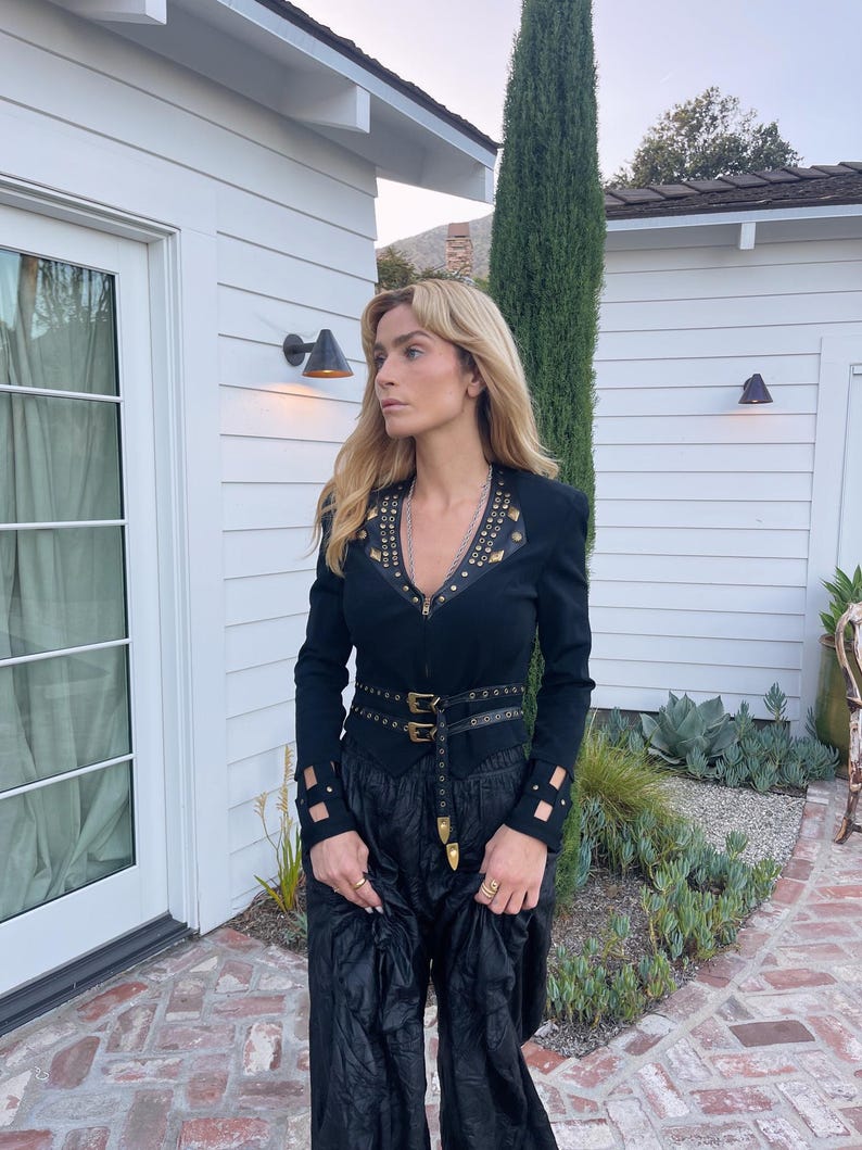 May include: Black long-sleeve top with gold-colored studs and a matching belt. The top has a zipper closure and cut-out details on the sleeves. The outfit is paired with black, wide-leg pants.