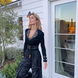 May include: A black long-sleeved top with gold embellishments and a matching belt, paired with black, gathered pants. The outfit is worn outdoors, with a white house and greenery in the background. The top has a V-neck and decorative details.