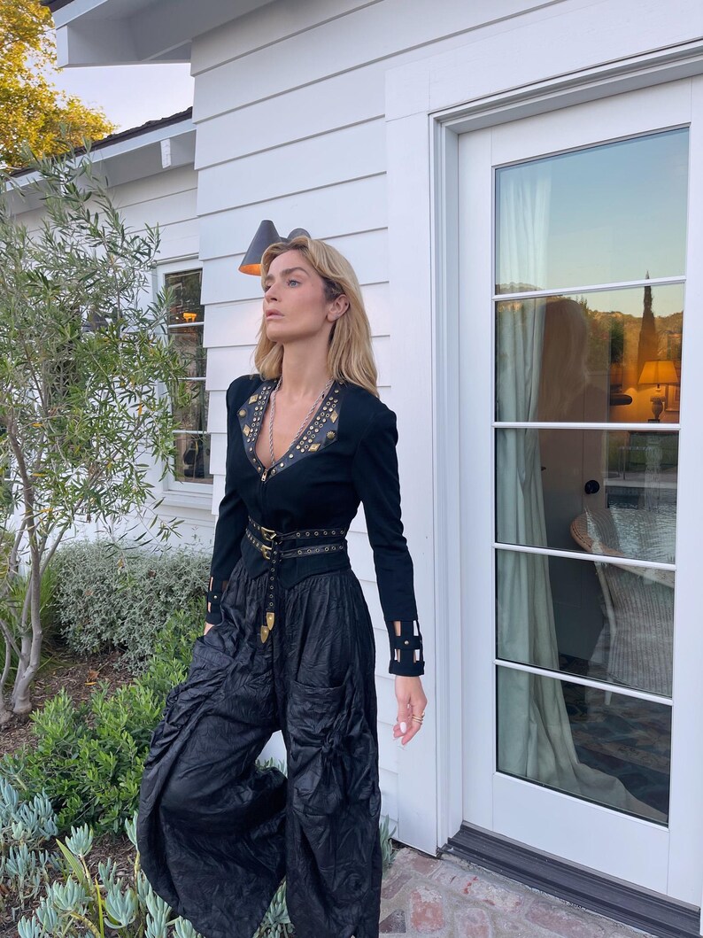 May include: A black long-sleeved top with gold embellishments and a matching belt, paired with black, gathered pants. The outfit is worn outdoors, with a white house and greenery in the background. The top has a V-neck and decorative details.