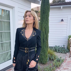 May include: Black jacket with gold-colored studs and a matching belt. The jacket has long sleeves with cut-out details and a V-neckline. The outfit includes a long black skirt with pockets. The model is standing outside.