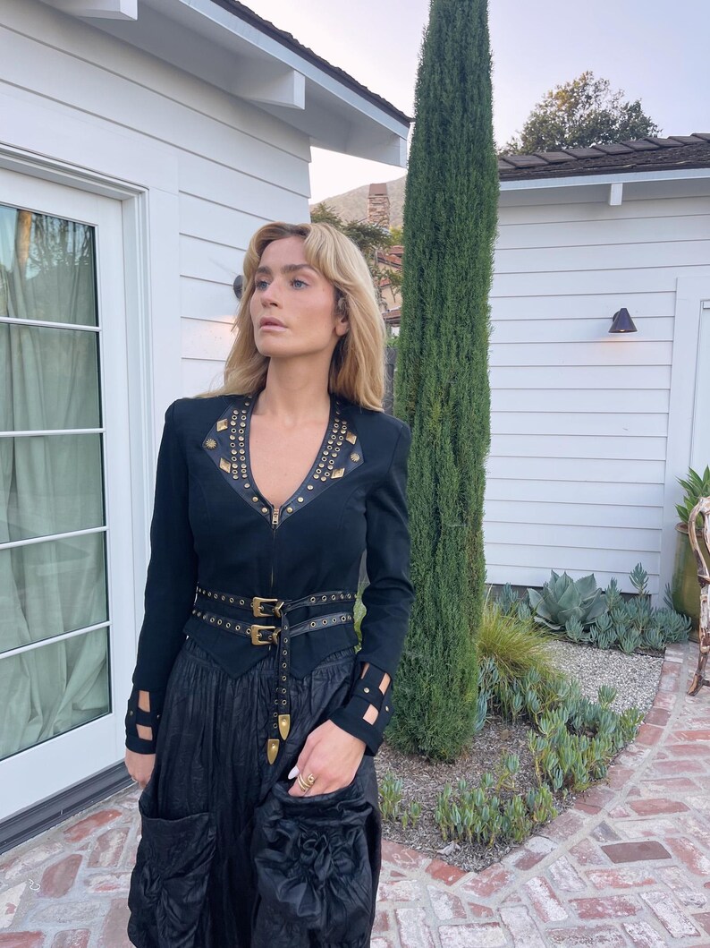 May include: Black jacket with gold-colored studs and a matching belt. The jacket has long sleeves with cut-out details and a V-neckline. The outfit includes a long black skirt with pockets. The model is standing outside.