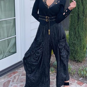 May include: A black outfit featuring a fitted jacket with gold-colored embellishments and a matching belt. The outfit includes wide-leg pants with gathered fabric details and black sandals. The ensemble is a stylish, edgy fashion statement.