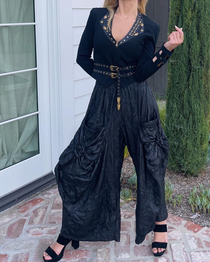 May include: A black outfit featuring a fitted jacket with gold-colored embellishments and a matching belt. The outfit includes wide-leg pants with gathered fabric details and black sandals. The ensemble is a stylish, edgy fashion statement.