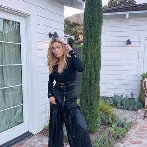 May include: A black long-sleeved top with gold-colored studs and a matching belt, paired with wide-leg black pants. The outfit is completed with black heeled sandals. The setting is outdoors, with a white building and greenery in the background.