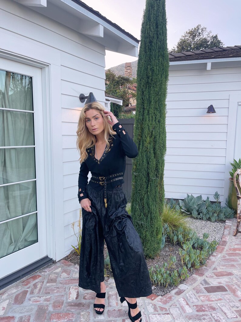 May include: A black long-sleeved top with gold-colored studs and a matching belt, paired with wide-leg black pants. The outfit is completed with black heeled sandals. The setting is outdoors, with a white building and greenery in the background.