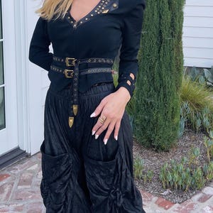 May include: Black jacket with gold-colored studs and a matching belt. The jacket has a V-neckline and long sleeves with cut-out details. The outfit includes black, gathered pants with large pockets, creating a unique, edgy look.