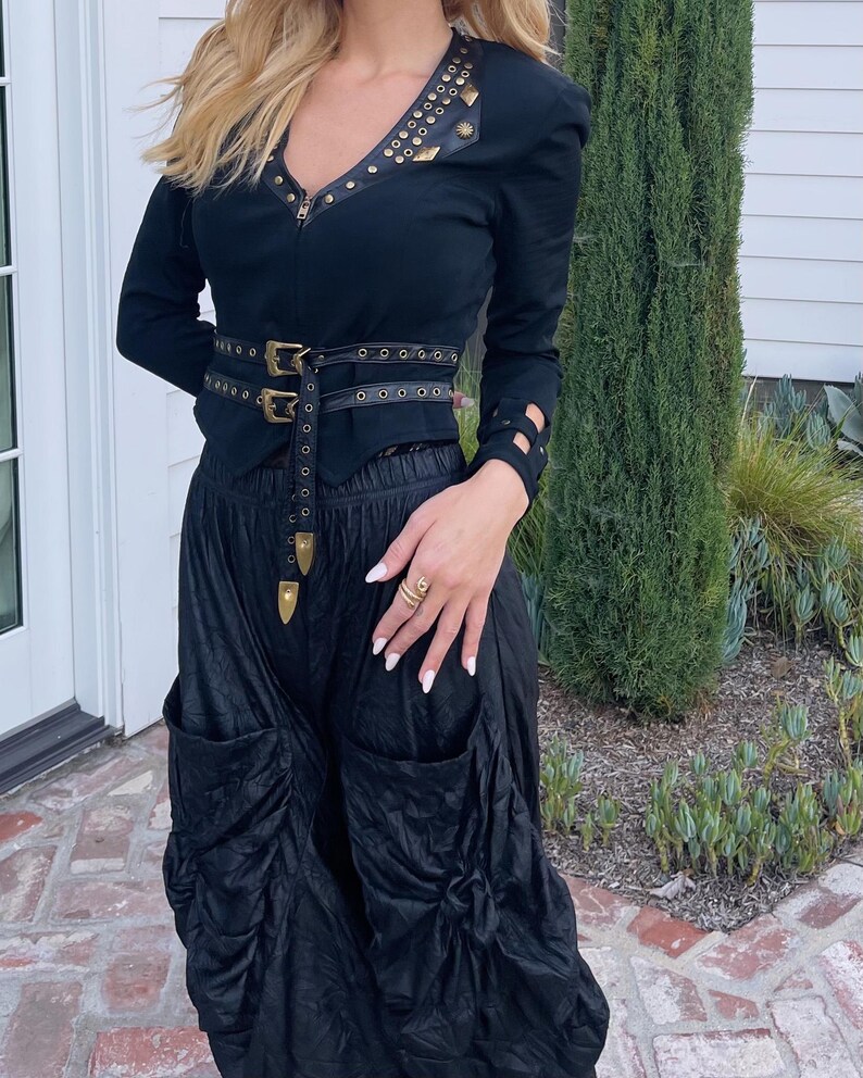 May include: Black jacket with gold-colored studs and a matching belt. The jacket has a V-neckline and long sleeves with cut-out details. The outfit includes black, gathered pants with large pockets, creating a unique, edgy look.