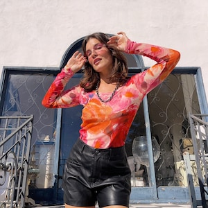 May include: A long-sleeved top with a vibrant floral pattern in shades of pink and orange, paired with black leather shorts. The top's design features a sheer fabric, and the shorts have a classic, tailored look. A necklace is also visible.